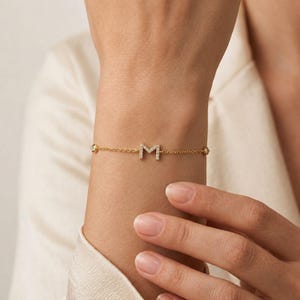 May include: A delicate gold chain bracelet featuring a central letter M pendant encrusted with small clear stones. The bracelet includes two additional small clear stones set along the chain on either side of the letter, creating a refined look.