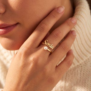 Peut inclure: Une bague ouverte dorée présentant le prénom Sonia écrit en lettres cursives à une extrémité et une petite perle blanche ronde à l'autre. La bague possède une finition métallique polie et une structure de bande ouverte ajustable.