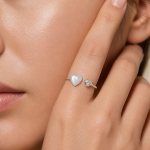 May include: A silver-toned open ring featuring two heart-shaped accents. One side displays a larger heart with a smooth, iridescent white inlay, while the other side features a smaller, clear, faceted heart-shaped stone set in metal.