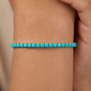 May include: A gold-toned tennis bracelet featuring a continuous row of round, vibrant turquoise-coloured stones set in a metallic frame. The bracelet encircles the wrist, showcasing a uniform pattern of bright blue circular gems held by gold prongs.