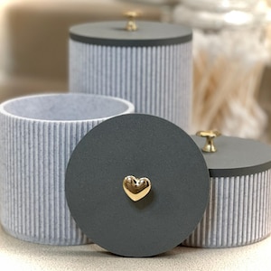 May include: Three cylindrical storage containers with a light gray ribbed texture. Each container has a dark gray circular lid topped with a small gold-colored knob. One lid features a gold-colored heart-shaped accent in the center.