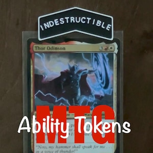 May include: A rectangular trading card featuring an illustration of Thor Odinson with lightning effects, encased in a clear plastic sleeve. A black, chevron-shaped topper with the white text INDESTRUCTIBLE is attached to the top of the card sleeve.