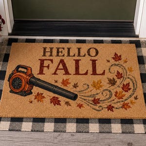 May include: A rectangular tan coir doormat featuring the text HELLO FALL in dark brown letters. An orange leaf blower is printed on the left side, with swirling autumn leaves in shades of red, orange, and yellow trailing from the nozzle.