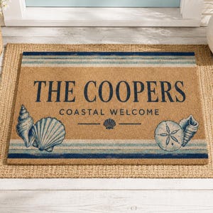 Könnte beinhalten: Eine rechteckige Fußmatte aus beigefarbenen Kokosfasern mit dunkelblauen horizontalen Streifen oben und unten. Der Text THE COOPERS COASTAL WELCOME ist in Dunkelblau gedruckt, ergänzt durch Abbildungen verschiedener Muscheln und eines Sanddollars.