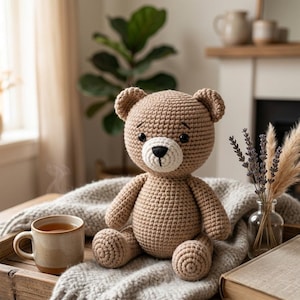 May include: A handmade, light brown crochet teddy bear with a white muzzle and black eyes sits on a gray blanket. A mug of tea and a small vase of dried flowers are nearby, creating a cozy scene. The bear is a soft toy.