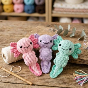 May include: Three handmade crochet axolotls in pink, purple, and mint green, each with a smiling face and frilly gills. A wooden crochet hook, a ball of white yarn, and a small jar with heart-shaped decorations are also visible.