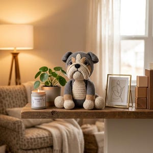 Peut inclure: Un bouledogue crocheté à la main, gris, beige et blanc, est assis sur une étagère en bois. À côté, une plante en pot, une bougie avec l'inscription "Lavender oil" et une impression d'art en ligne encadrée. La scène se déroule dans une pièce chaleureuse et bien éclairée.