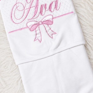 May include: White baby blanket with pink embroidered details. The name "Ava" is displayed in a cursive font above a pink bow. The top section has a quilted pattern, while the bottom is a solid white.