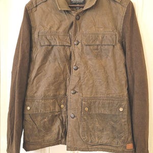 May include: An olive green jacket with a button-up front, featuring a collar, shoulder epaulets, two chest pockets with flaps, and two large lower pockets with snap closures. The brand label reads Scotch & Soda, Amsterdam Couture.