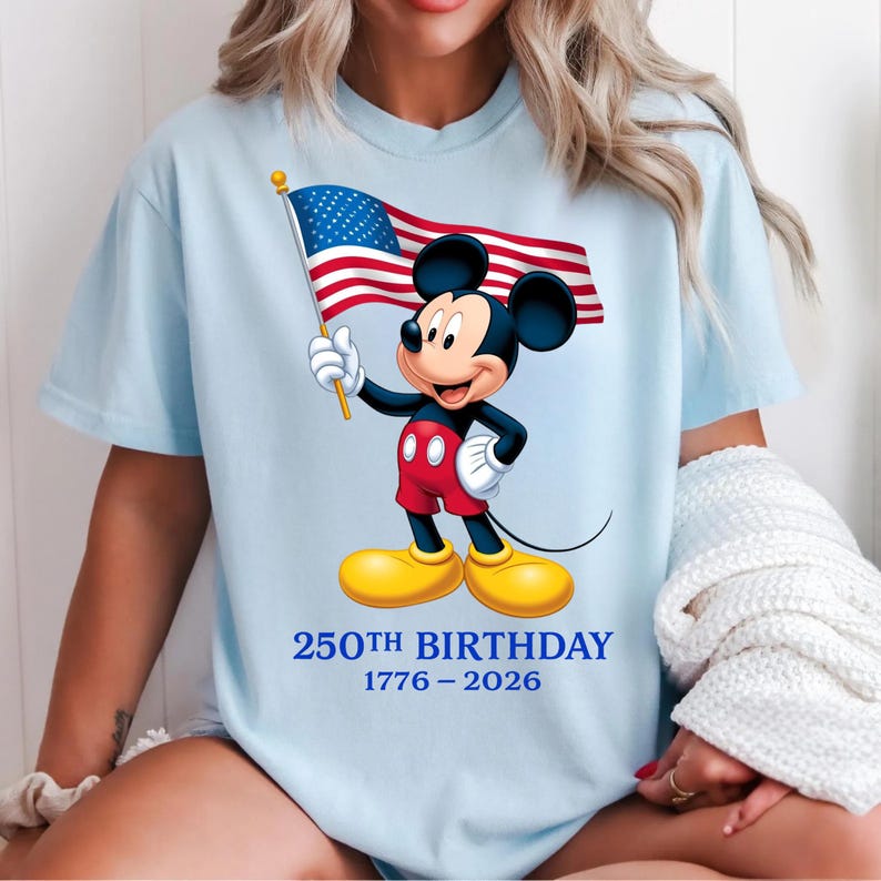 Puede incluir: Camiseta azul claro con Mickey Mouse sosteniendo una bandera estadounidense. Mickey lleva pantalones cortos rojos, zapatos amarillos y guantes blancos. El texto "250TH BIRTHDAY 1776-2026" est&aacute; impreso debajo. Estilo casual.