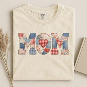 May include: Cream-colored t-shirt with the word "MOM" in a patchwork design. The letters are made of denim, floral, and gingham fabrics, with a red heart in the center of the "O". The shirt has a crew neck and a Comfort Colors tag.