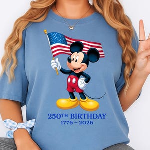 Puede incluir: Camiseta azul claro con Mickey Mouse sosteniendo una bandera estadounidense. Mickey lleva pantalones cortos rojos, guantes blancos y zapatos amarillos. El texto "250th BIRTHDAY 1776-2026" est&aacute; impreso debajo en azul.