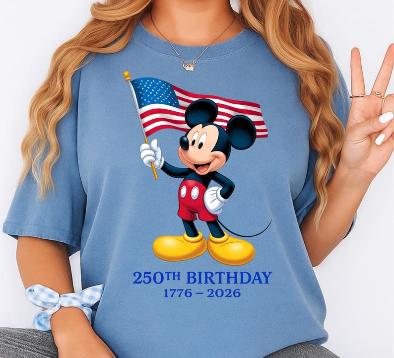 Puede incluir: Camiseta azul claro con Mickey Mouse sosteniendo una bandera estadounidense. Mickey lleva pantalones cortos rojos, guantes blancos y zapatos amarillos. El texto "250th BIRTHDAY 1776-2026" est&aacute; impreso debajo en azul.