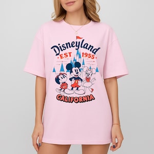 Puede incluir: Camiseta rosa claro con un dise&ntilde;o de Disneyland. El dise&ntilde;o incluye a Mickey Mouse, Minnie Mouse y Goofy, con las palabras "Disneyland EST 1955 CALIFORNIA" en una fuente divertida. La camiseta tiene mangas cortas y cuello redondo.