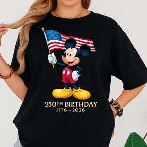 Puede incluir: Camiseta negra con un dise&ntilde;o de Mickey Mouse sosteniendo una bandera estadounidense. Mickey lleva pantalones cortos rojos y zapatos amarillos. El texto "250th BIRTHDAY 1776-2026" est&aacute; impreso debajo.