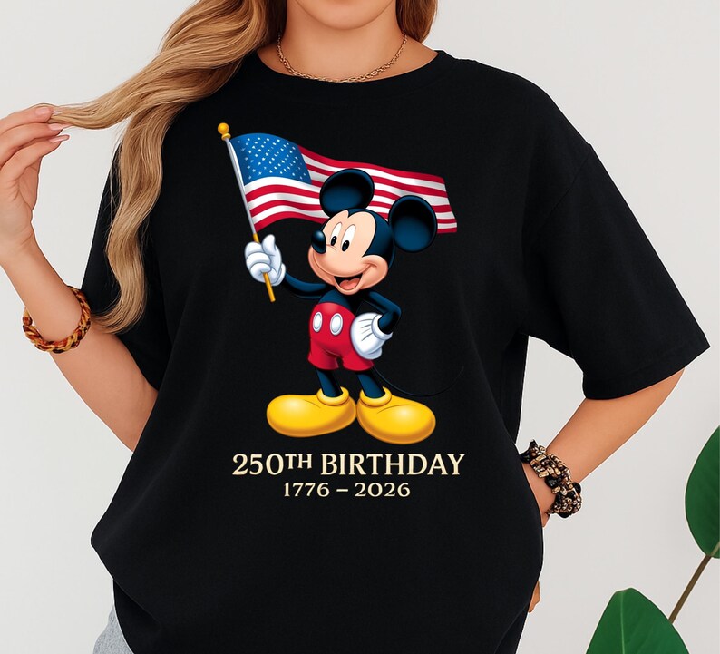 Puede incluir: Camiseta negra con un dise&ntilde;o de Mickey Mouse sosteniendo una bandera estadounidense. Mickey lleva pantalones cortos rojos y zapatos amarillos. El texto "250th BIRTHDAY 1776-2026" est&aacute; impreso debajo.