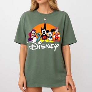 Pu&ograve; includere: Una maglietta verde oliva con una grafica Disney. La grafica presenta personaggi dei cartoni animati davanti a un sole arancione, con la scritta "Disney" in bianco sotto. La maglietta ha maniche corte e girocollo.