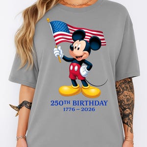 Puede incluir: Camiseta gris con un dibujo de Mickey Mouse sosteniendo una bandera estadounidense. Mickey lleva pantalones cortos rojos, guantes blancos y zapatos amarillos. El texto "250th BIRTHDAY 1776-2026" est&aacute; impreso en azul debajo.