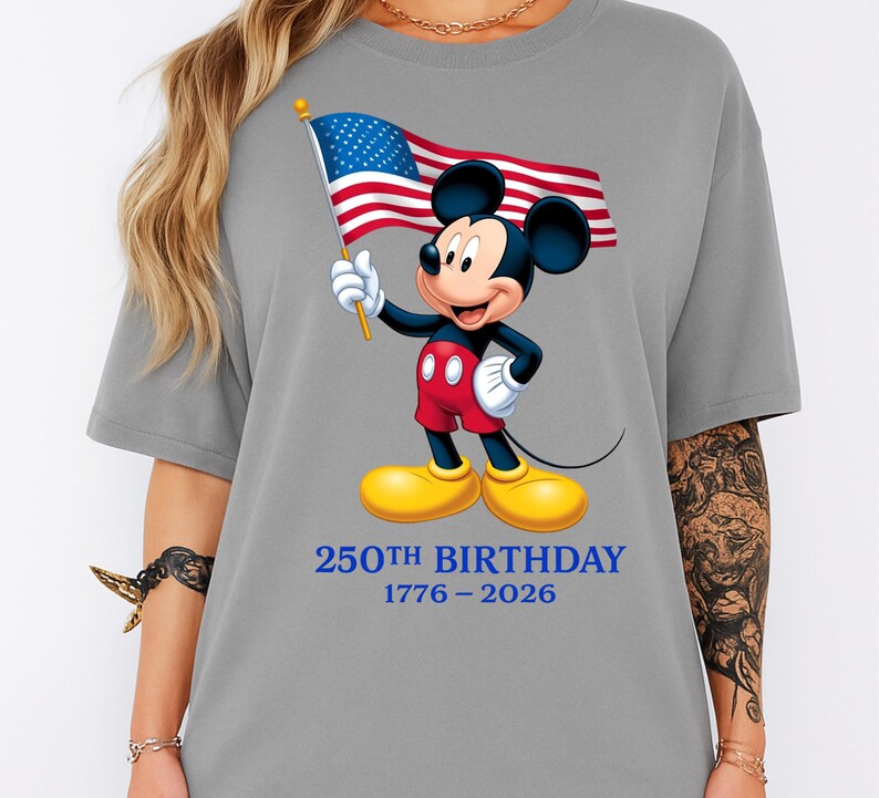 Puede incluir: Camiseta gris con un dibujo de Mickey Mouse sosteniendo una bandera estadounidense. Mickey lleva pantalones cortos rojos, guantes blancos y zapatos amarillos. El texto "250th BIRTHDAY 1776-2026" est&aacute; impreso en azul debajo.