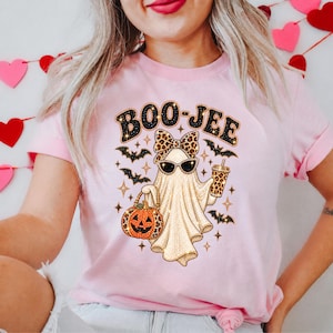 May include: A light pink t-shirt featuring a graphic of a ghost wearing a leopard print bow and sunglasses. The ghost holds a leopard print pumpkin bucket and a drink cup. The text BOO-JEE is printed above, surrounded by black bats and gold stars.