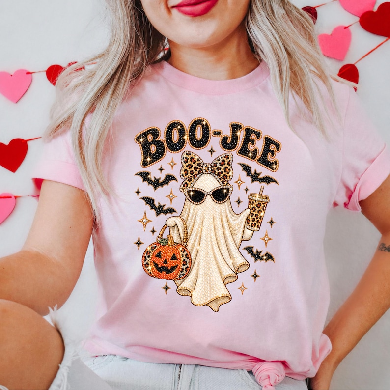 May include: A light pink t-shirt featuring a graphic of a ghost wearing a leopard print bow and sunglasses. The ghost holds a leopard print pumpkin bucket and a drink cup. The text BOO-JEE is printed above, surrounded by black bats and gold stars.