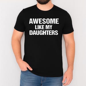 K&ouml;nnte beinhalten: Schwarzes T-Shirt mit Rundhalsausschnitt und dem wei&szlig;en Text "AWESOME LIKE MY DAUGHTERS". Das Shirt besteht aus einem weichen Material und eignet sich f&uuml;r Freizeitkleidung. Der Text ist mittig auf der Brust platziert.