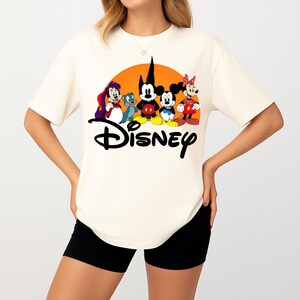 Pu&ograve; includere: T-shirt color crema con grafica Disney. Il design include Topolino, Minnie e altri personaggi davanti a un sole arancione. La parola "Disney" &egrave; scritta in nero sotto i personaggi.