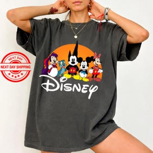 Pu&ograve; includere: T-shirt grigio scuro con una grafica Disney con Topolino, Minnie e Pippo, su sfondo arancione. La parola "Disney" &egrave; scritta in bianco sotto i personaggi.