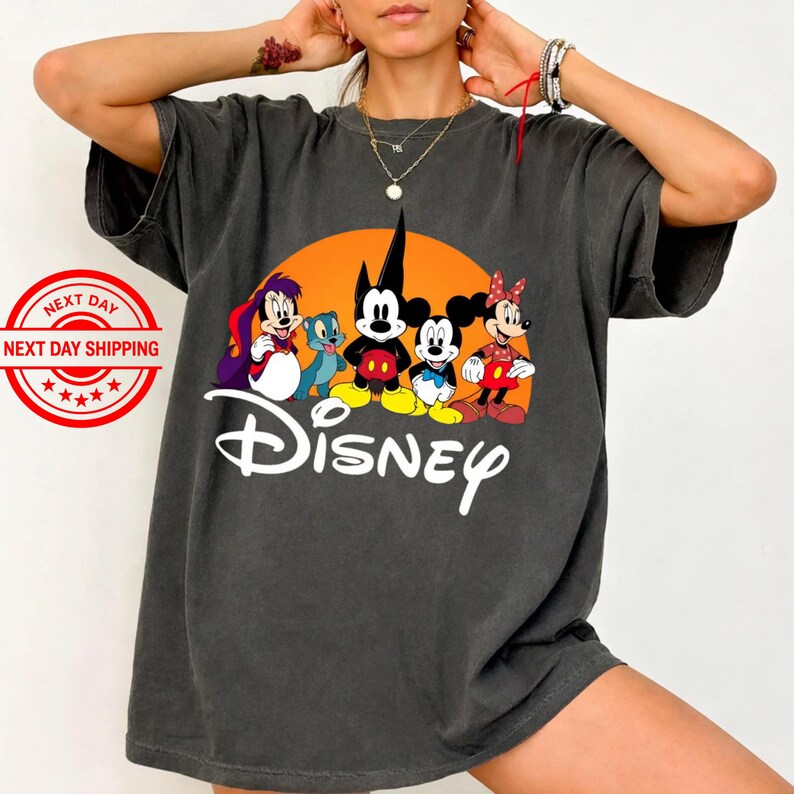 Pu&ograve; includere: T-shirt grigio scuro con una grafica Disney con Topolino, Minnie e Pippo, su sfondo arancione. La parola "Disney" &egrave; scritta in bianco sotto i personaggi.