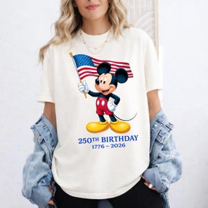 Puede incluir: Camiseta color crema con Mickey Mouse sosteniendo una bandera estadounidense. Mickey lleva pantalones cortos rojos, guantes blancos y zapatos amarillos. El texto "250TH BIRTHAY 1776-2026" est&aacute; impreso en azul debajo.