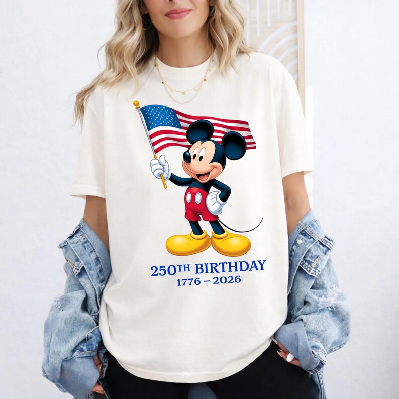 Puede incluir: Camiseta color crema con Mickey Mouse sosteniendo una bandera estadounidense. Mickey lleva pantalones cortos rojos, guantes blancos y zapatos amarillos. El texto "250TH BIRTHAY 1776-2026" est&aacute; impreso en azul debajo.