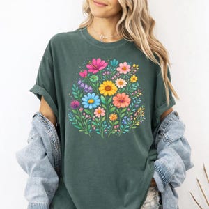 May include: Dark green t-shirt featuring a vibrant floral design. The graphic showcases a bouquet of colorful flowers, including pink, yellow, blue, and orange blooms, surrounded by green leaves. The shirt has short sleeves and a relaxed fit.
