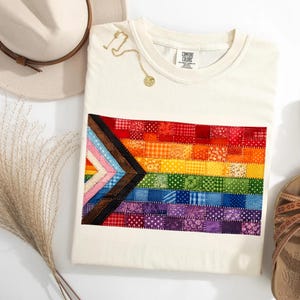 May include: A cream-colored t-shirt featuring a rectangular graphic of the Progress Pride flag composed of small, multi-patterned fabric squares in red, orange, yellow, green, blue, purple, brown, black, light blue, pink, and white.