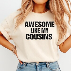K&ouml;nnte beinhalten: Ein cremefarbenes T-Shirt mit dem Text "AWESOME LIKE MY COUSINS" in fetten, schwarzen Buchstaben. Das Shirt hat kurze &Auml;rmel und wird mit einer Jeans getragen.