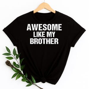 K&ouml;nnte beinhalten: Schwarzes T-Shirt mit dem wei&szlig;en Text "AWESOME LIKE MY BROTHER". Das T-Shirt h&auml;ngt an einem Holzb&uuml;gel. Ein Zweig mit Gr&uuml;n befindet sich in der unteren linken Ecke.