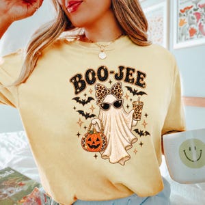 May include: A pale yellow t-shirt featuring a graphic of a ghost wearing a leopard print bow and sunglasses. The ghost holds a leopard print pumpkin bucket and a drink cup. The text BOO-JEE is printed above, surrounded by small black bats.