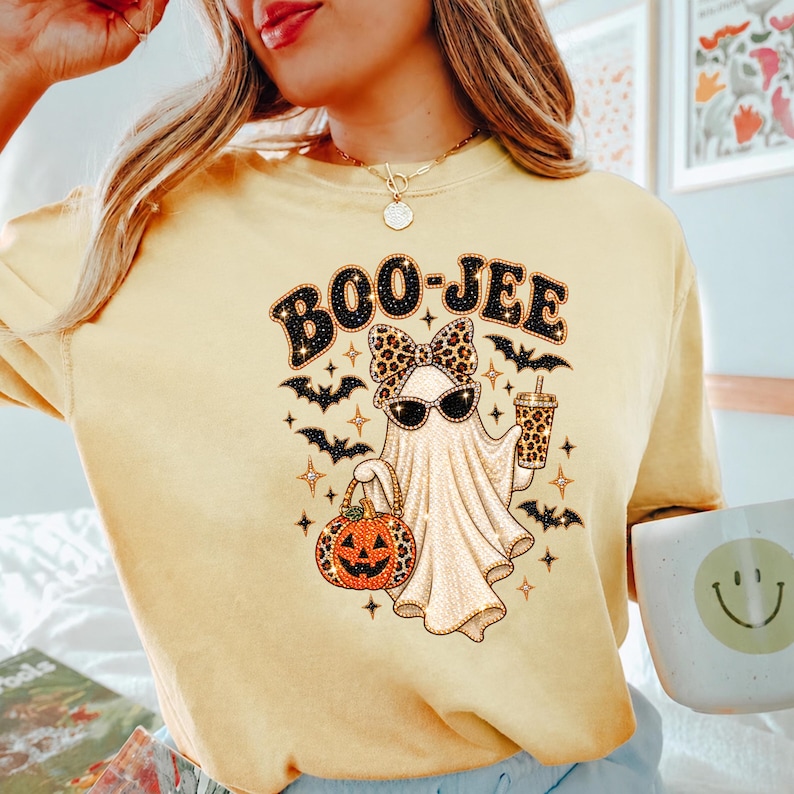 May include: A pale yellow t-shirt featuring a graphic of a ghost wearing a leopard print bow and sunglasses. The ghost holds a leopard print pumpkin bucket and a drink cup. The text BOO-JEE is printed above, surrounded by small black bats.