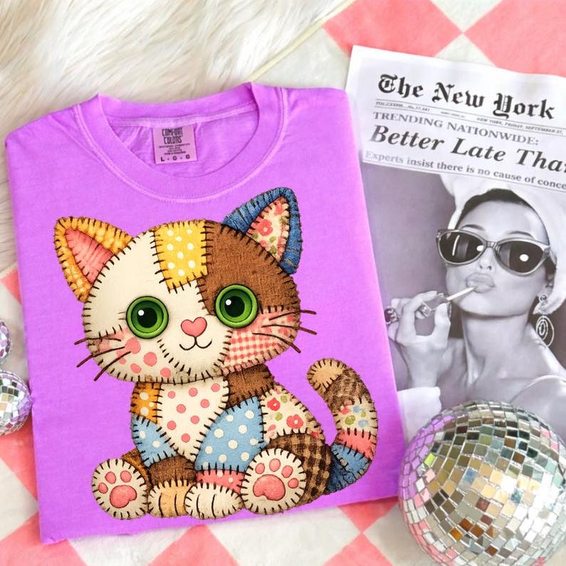 May include: A lilac-colored t-shirt featuring a patchwork cat design with green eyes, a pink nose, and various fabric patterns. The cat is sitting, and the shirt is displayed with a newspaper and a disco ball.