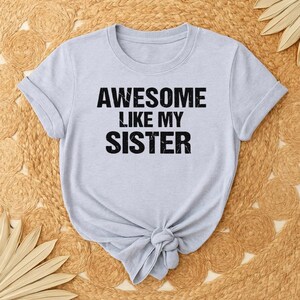 K&ouml;nnte beinhalten: Hellgraues T-Shirt mit dem Text "AWESOME LIKE MY SISTER" in fetten schwarzen Buchstaben. Das Shirt hat kurze &Auml;rmel und ist am unteren Saum geknotet. Es liegt auf einer gewebten, beigefarbenen Oberfl&auml;che.