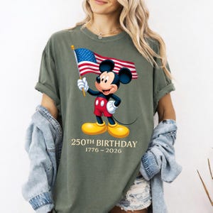 Puede incluir: Camiseta verde oliva con Mickey Mouse sosteniendo una bandera estadounidense. Mickey lleva pantalones cortos rojos y zapatos amarillos. El texto "250TH BIRTHDAY 1776-2026" est&aacute; impreso debajo.