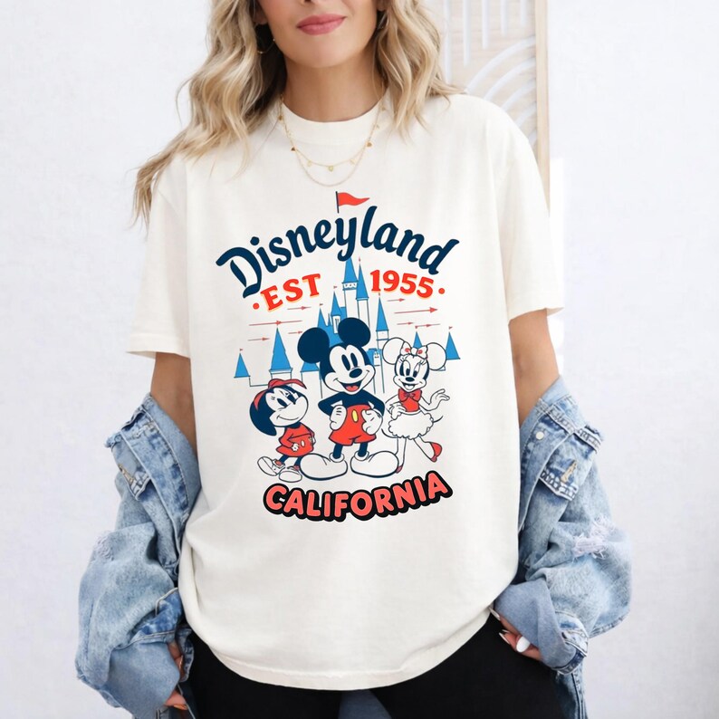 Puede incluir: Camiseta color crema con un gr&aacute;fico de Disneyland. El dise&ntilde;o incluye a Mickey Mouse, Minnie Mouse y Donald Duck, con el texto "Disneyland EST 1955 CALIFORNIA" en rojo y azul. La camiseta tiene mangas cortas y un corte holgado.