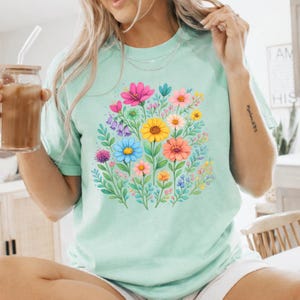 May include: A light green t-shirt featuring a vibrant floral design. The design includes a variety of colorful flowers in shades of pink, yellow, blue, and orange, surrounded by green leaves. The shirt has a round neckline and short sleeves.