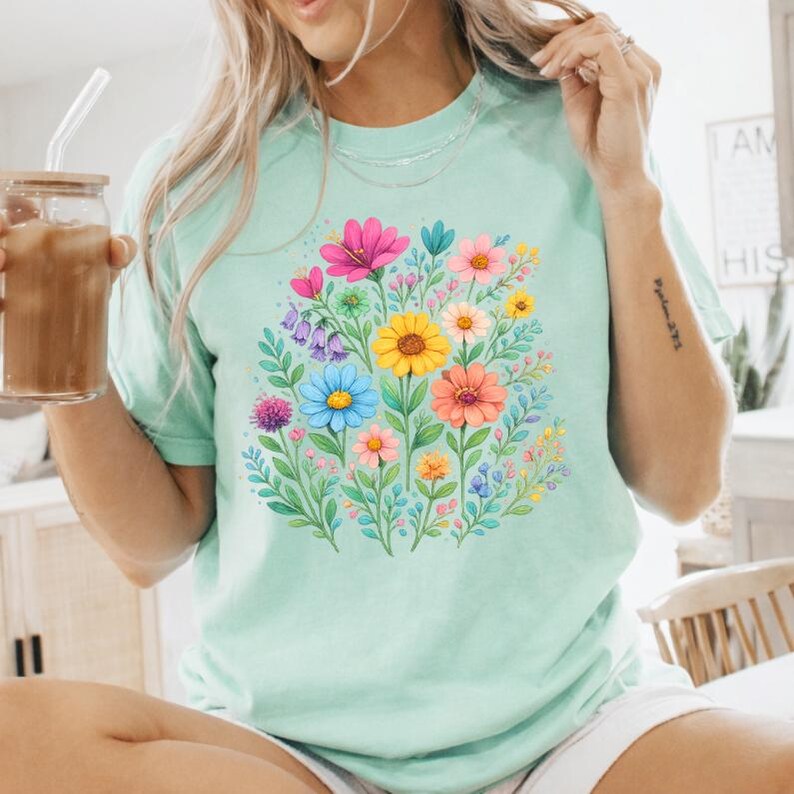 May include: A light green t-shirt featuring a vibrant floral design. The design includes a variety of colorful flowers in shades of pink, yellow, blue, and orange, surrounded by green leaves. The shirt has a round neckline and short sleeves.