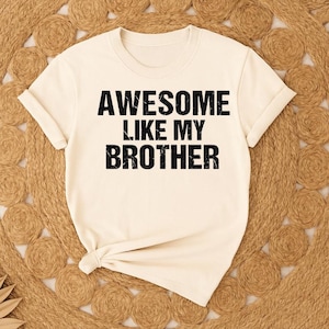 K&ouml;nnte beinhalten: Cremefarbenes T-Shirt mit dem Text "AWESOME LIKE MY BROTHER" in fetten, schwarzen Buchstaben. Das Shirt hat kurze &Auml;rmel und einen runden Ausschnitt. Das Shirt liegt auf einer gewebten, hellbraunen Oberfl&auml;che.