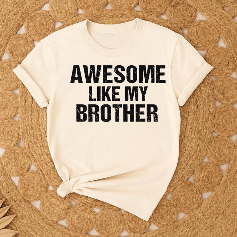 K&ouml;nnte beinhalten: Cremefarbenes T-Shirt mit dem Text "AWESOME LIKE MY BROTHER" in fetten, schwarzen Buchstaben. Das Shirt hat kurze &Auml;rmel und einen runden Ausschnitt. Das Shirt liegt auf einer gewebten, hellbraunen Oberfl&auml;che.