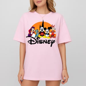Pu&ograve; includere: T-shirt rosa chiaro con grafica Disney. La grafica include Topolino, Minnie e altri personaggi Disney davanti a un sole arancione. La parola "Disney" &egrave; scritta in nero sotto i personaggi.