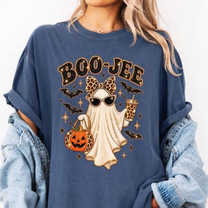 May include: A blue short-sleeve t-shirt featuring a graphic of a ghost wearing leopard print sunglasses and a matching bow. The ghost holds a pumpkin bucket and a leopard print cup. The text BOO-JEE is printed above with small bats and stars.