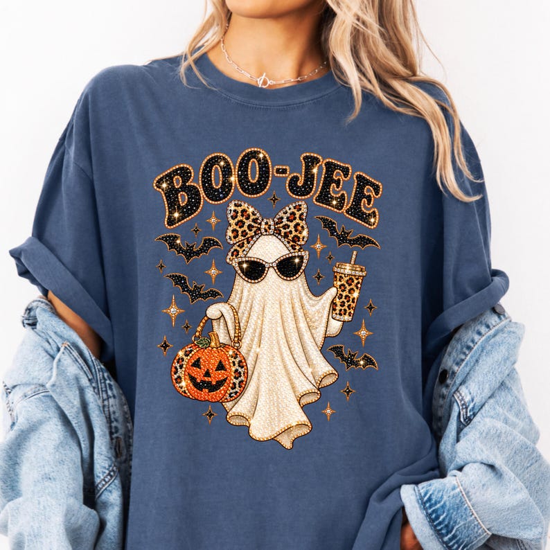 May include: A blue short-sleeve t-shirt featuring a graphic of a ghost wearing leopard print sunglasses and a matching bow. The ghost holds a pumpkin bucket and a leopard print cup. The text BOO-JEE is printed above with small bats and stars.