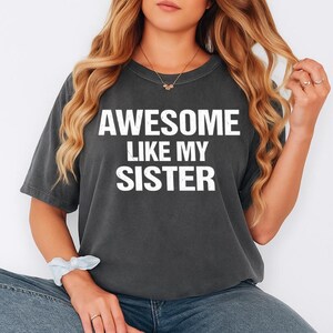 K&ouml;nnte beinhalten: Dunkelgraues T-Shirt mit dem wei&szlig;en Text "AWESOME LIKE MY SISTER". Das T-Shirt hat einen Rundhalsausschnitt. Eine Person tr&auml;gt das T-Shirt und Jeans.