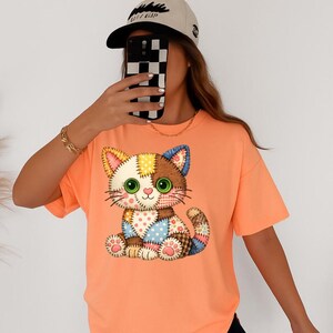 May include: Orange short-sleeved t-shirt featuring a patchwork cat design. The cat has green eyes, a pink heart nose, and a variety of colorful patches. The shirt is a solid orange color.